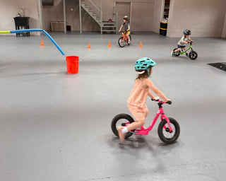 Children riding balance bikes in a large indoor space with wooden beams.