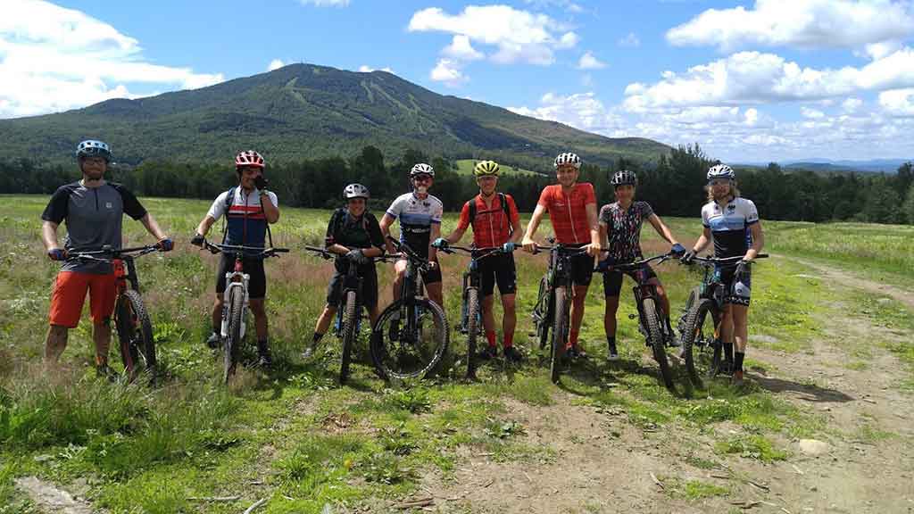 A group of eight mountain bikers in helmets and jerseys lined up with their bikes in a meadow at Darling Hill, with Burke Mountain rising under a clear blue sky behind them