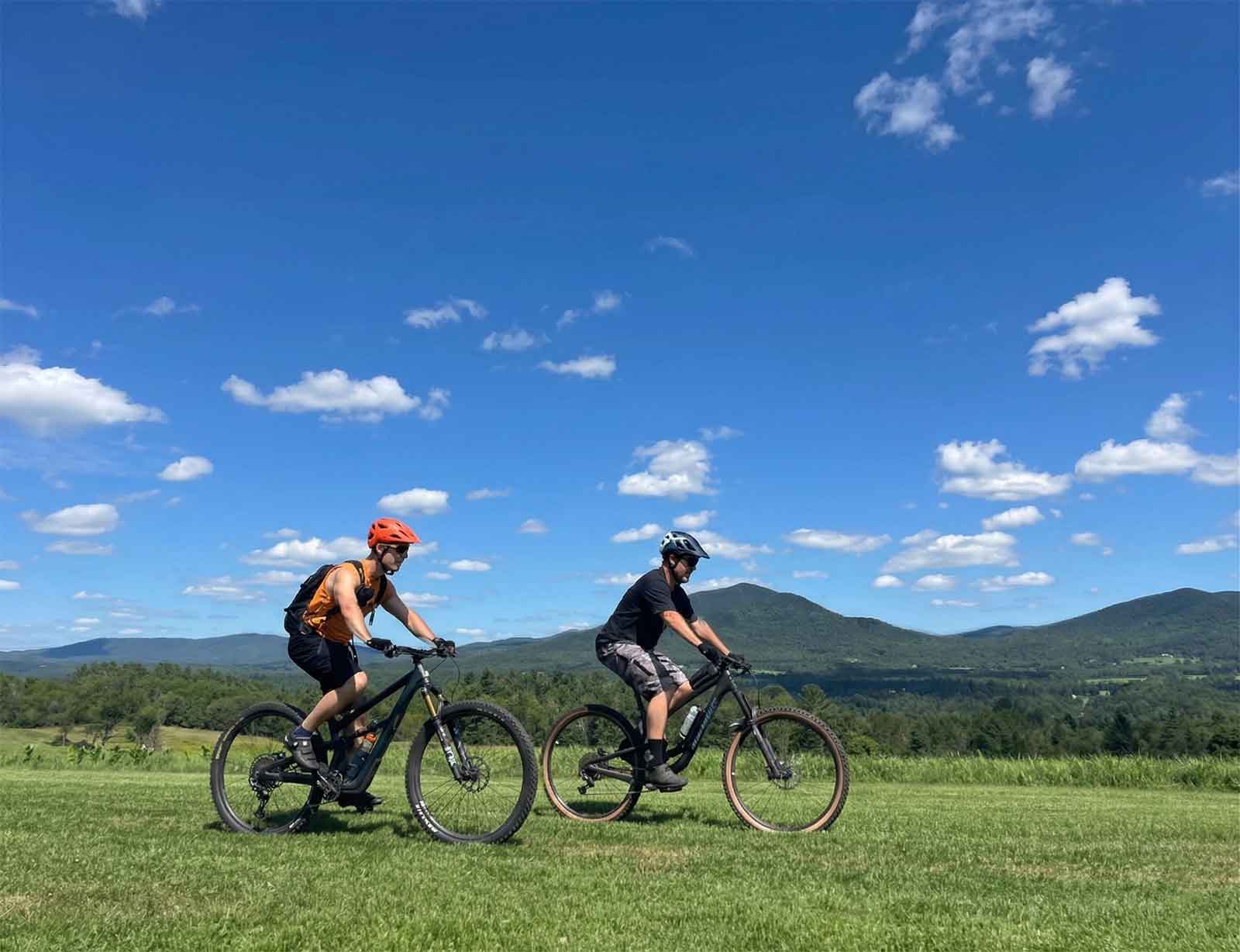 Two mountain bikers riding side by side across an open grassy hilltop in the Litchfield Hills, with mountain ridges and a bright blue sky in the distance