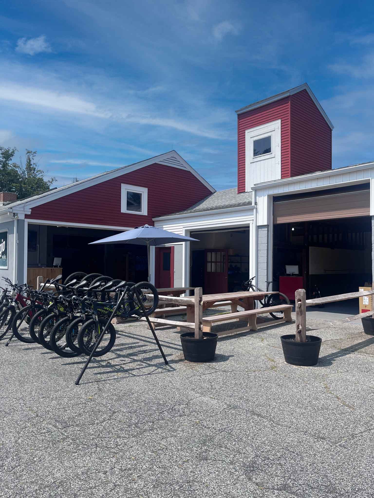 The red-barn Rifugio Litchfield Hills shop on Bantam Road, with a row of mountain bikes lined up out front, picnic tables under a blue umbrella, and open garage bays revealing the workshop inside