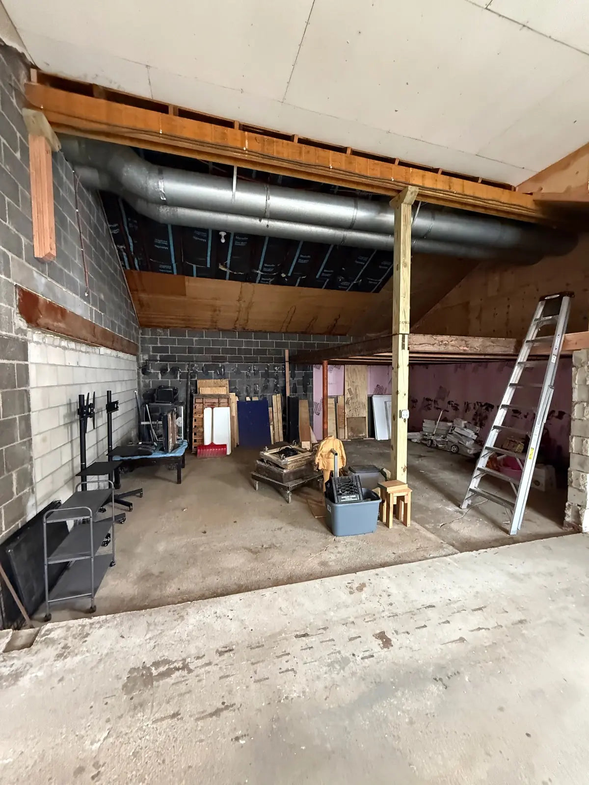 A cluttered storage bay tucked under the roof framing, with cinderblock walls, ductwork overhead, and a ladder and rolling carts on the bare concrete floor