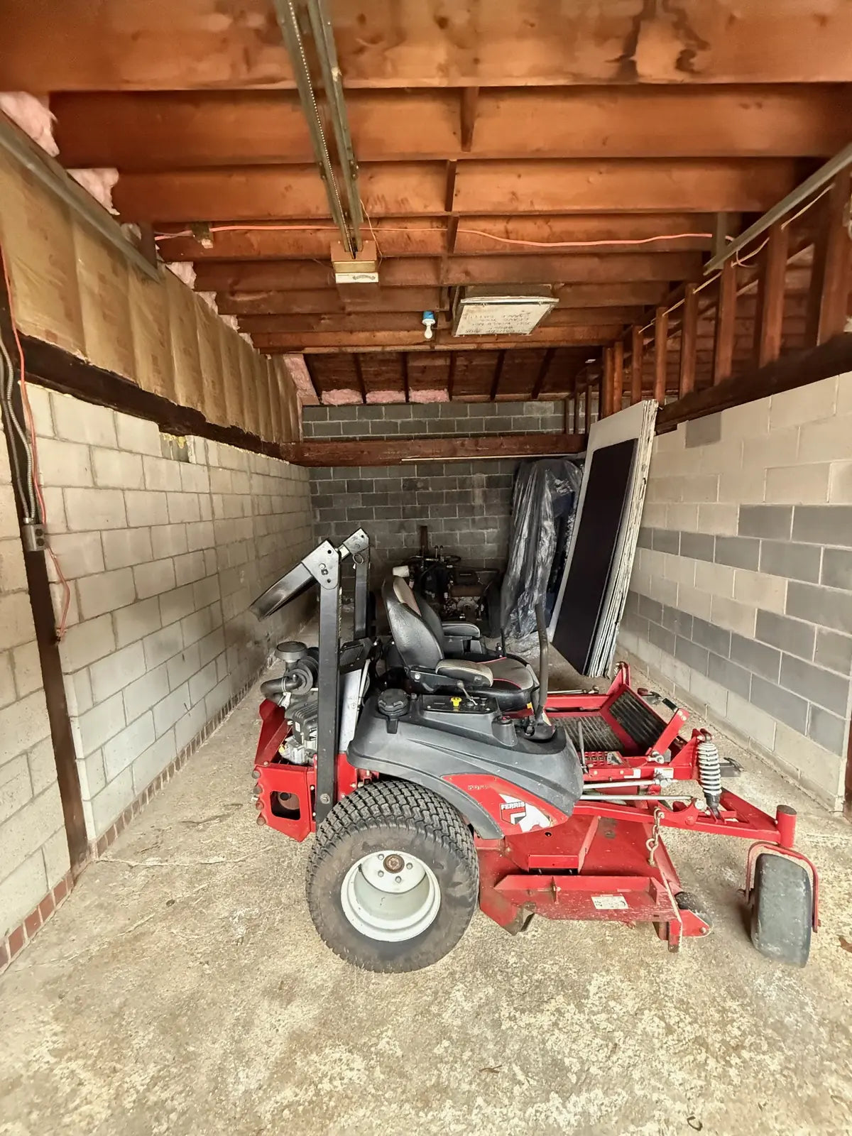 A narrow storage bay with rough wood ceiling beams and cinderblock walls, a red riding mower parked on the concrete floor