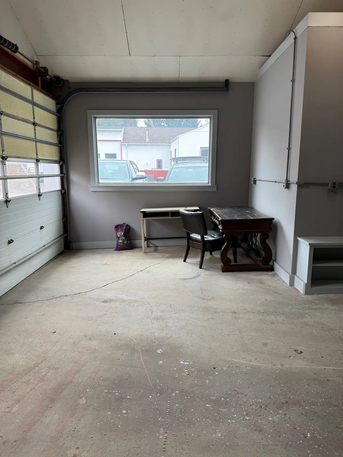 A gutted room freshly painted grey, with a picture window, an old desk and office chair, and a bag of supplies left on the floor