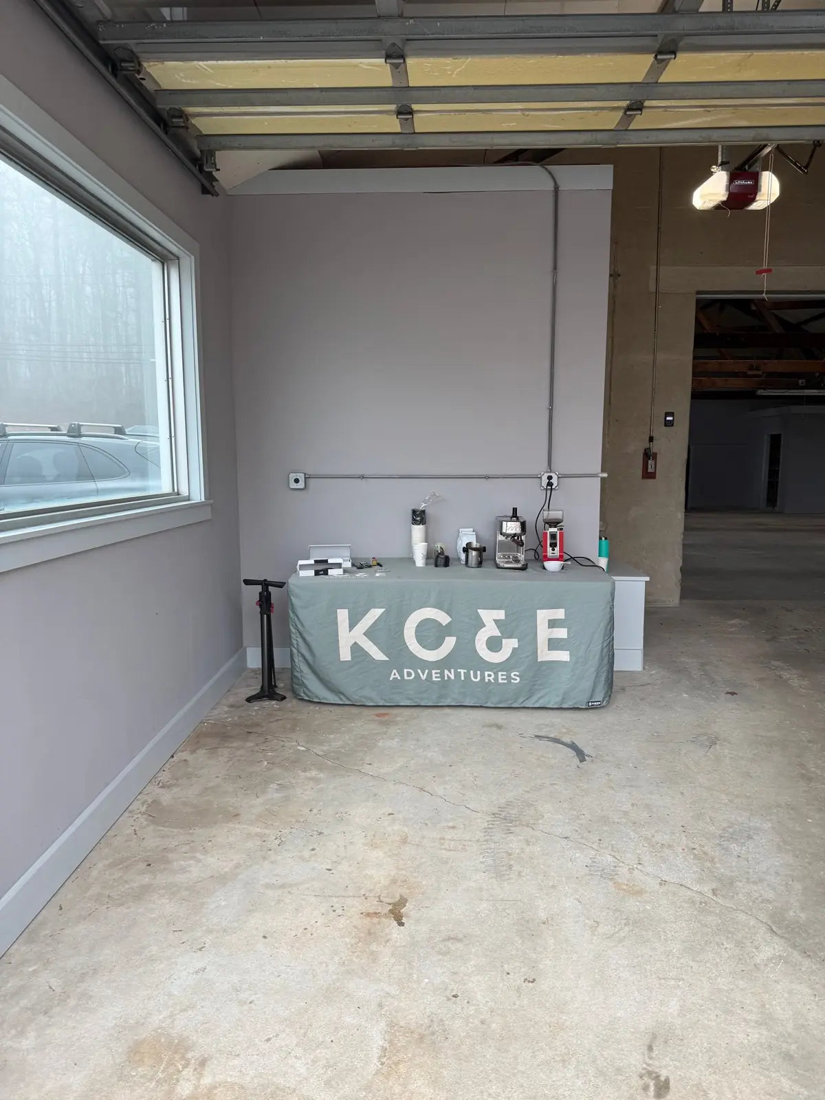 A folding table skirted in a KC&E Adventures cloth, set up with an espresso machine, grinder, and paper cups, with a bike floor pump leaning against the wall beside it