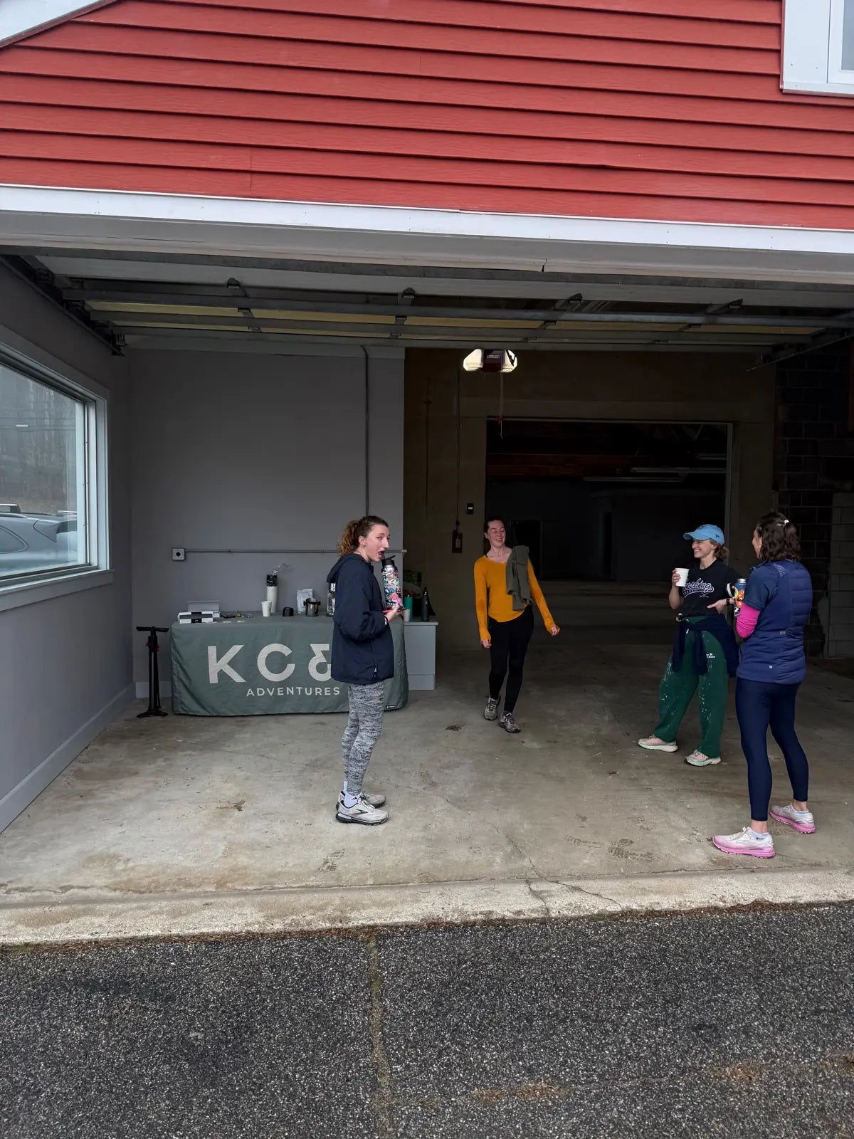 Four people standing on the concrete apron outside the open garage door, several holding coffee cups, the KC&E Adventures coffee table visible just inside